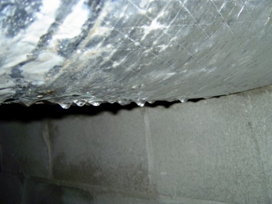 Water droplets form and drip from a pipe covered in condensation above a concrete block wall, suggesting moisture accumulation or a possible leak.